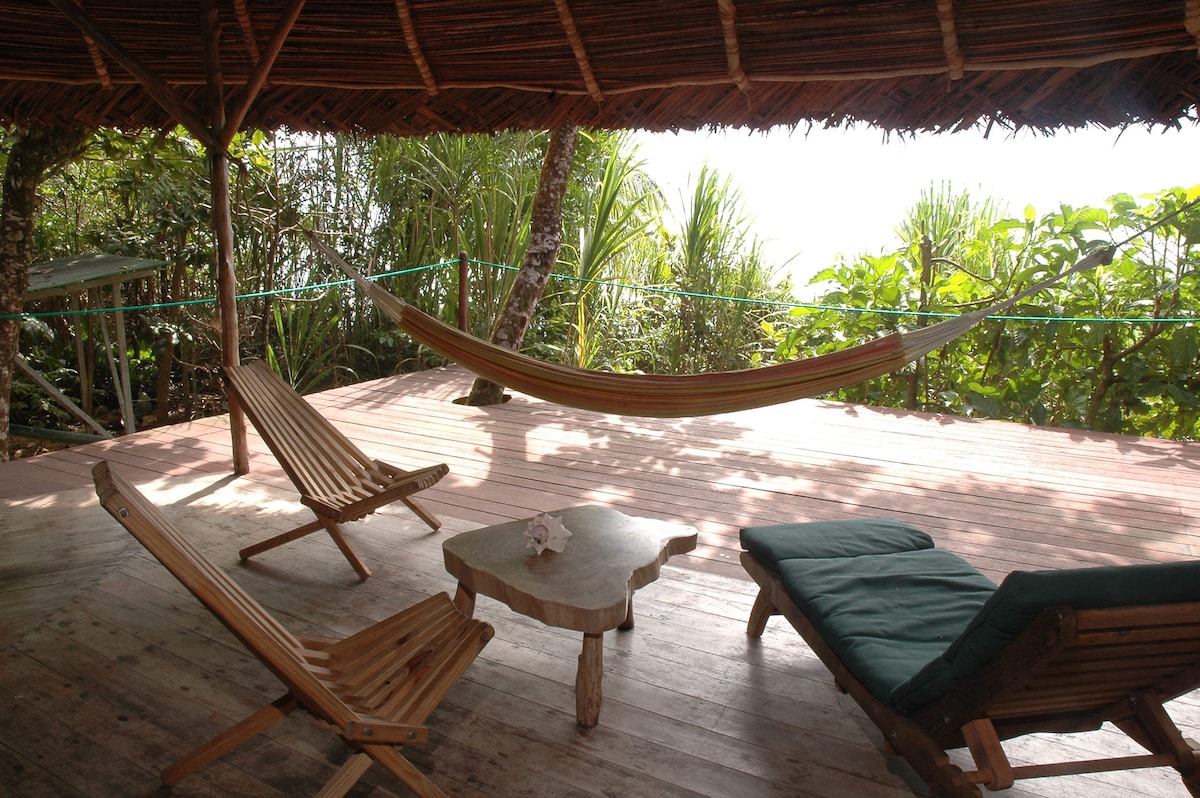 A spacious wooden terrace is featured, showcasing a hammock gently hanging between two trees. Two lounge chairs and a small coffee table create a relaxing space, while lush tropical greenery frames the scene, providing a serene and inviting environment.