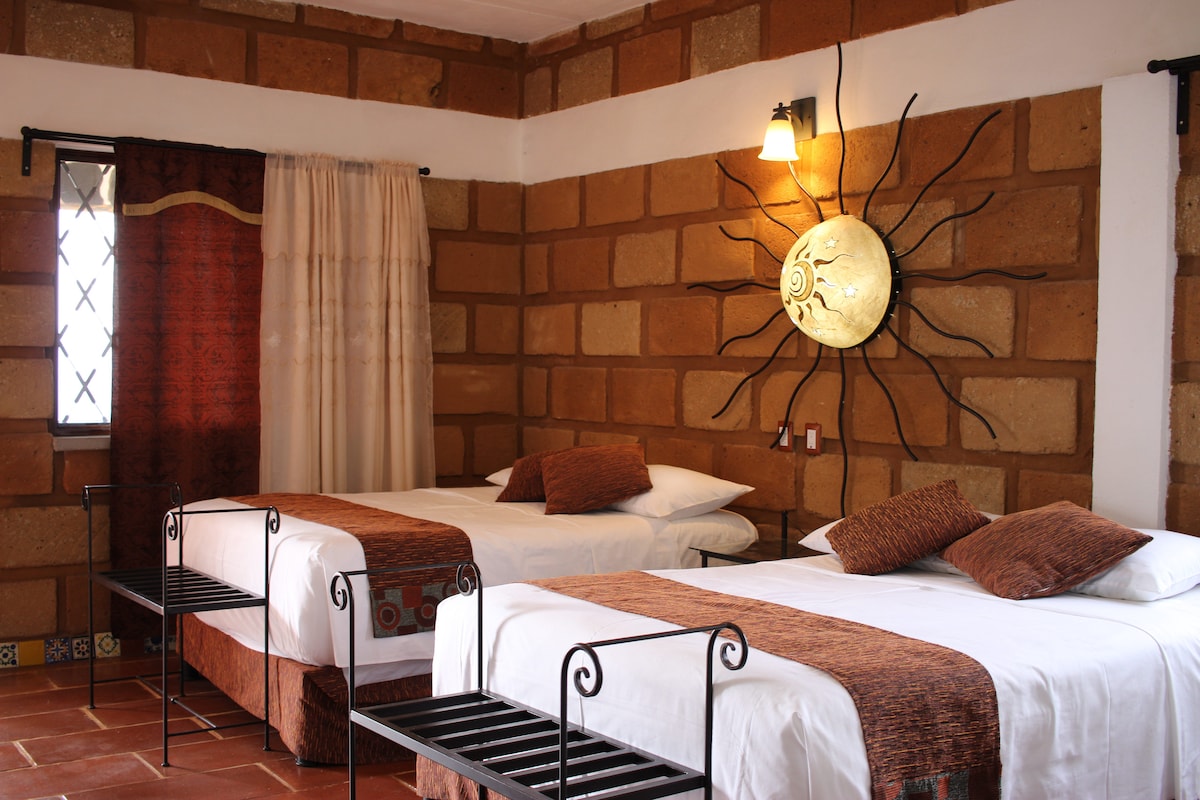 A room featuring two spacious beds with decorative brown and orange accents. The walls are crafted from natural stone, and a sun-themed wall hanging draws attention. A window with sheer curtains allows natural light to enter, complemented by a wall lamp providing additional illumination.