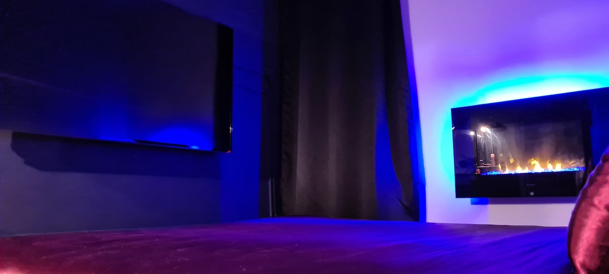 A spacious bed is centered in the room, accompanied by a wall-mounted television on one side. A modern electric fireplace is positioned nearby, illuminated with blue tones, creating an ambient setting. Soft drapes cover the window, contributing to the overall cozy atmosphere.