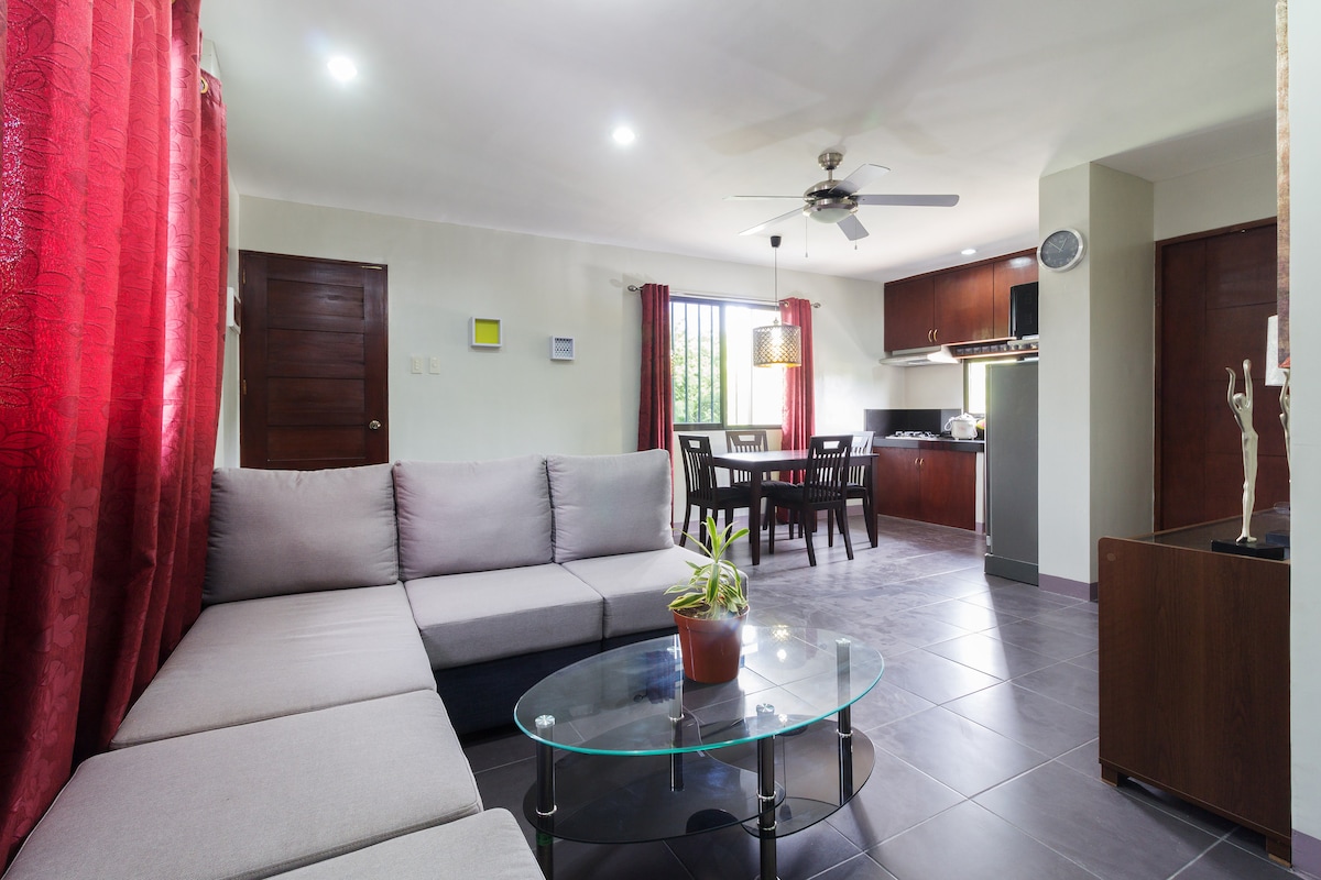 A spacious living area features a light grey sectional sofa surrounding a round glass coffee table, complemented by a small indoor plant. Bright red curtains frame large windows, allowing natural light to illuminate the open space, which leads to a dining area and a fully equipped kitchen.