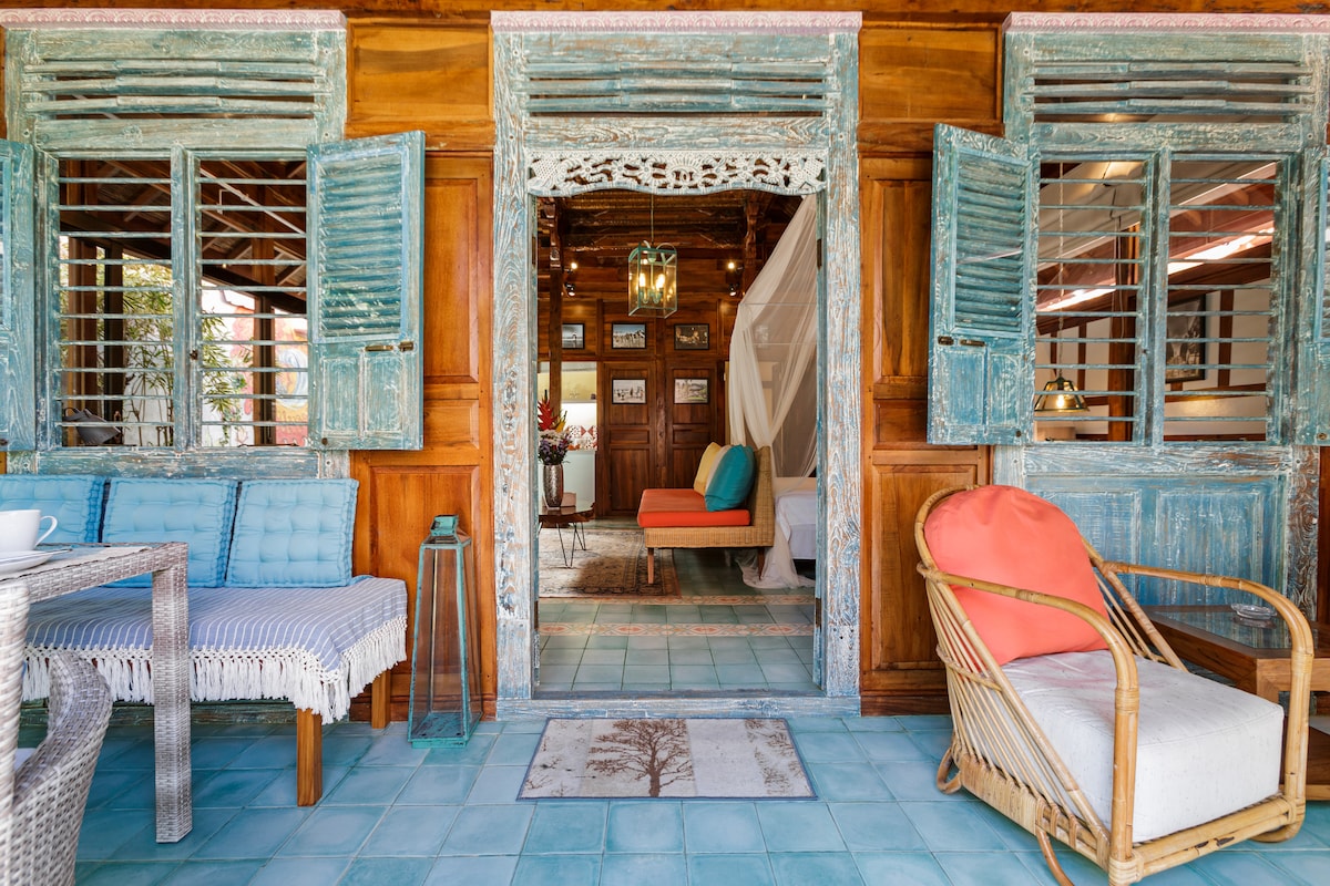 The entrance to the villa features wooden shutters painted in blue, providing a rustic charm. A bamboo-framed armchair complements a small table with a cup, while the spacious interior is visible beyond. Colorful cushions add a welcoming touch to the space.