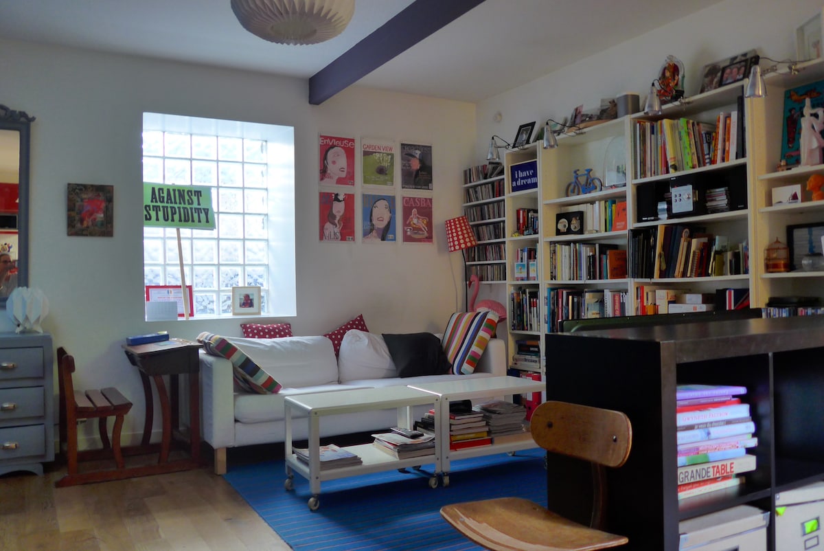 A bright living area features a comfortable white couch adorned with colorful throw pillows. A collection of books fills the shelves against the wall, alongside framed artwork. A blue rug adds a touch of color to the wooden floor, and a side table is visible near the window.