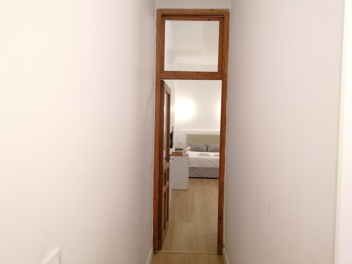 A narrow hallway is presented, featuring white walls and a wooden door at the end. Light floods the area, hinting at an inviting living space beyond the doorway, where a bed is faintly visible.