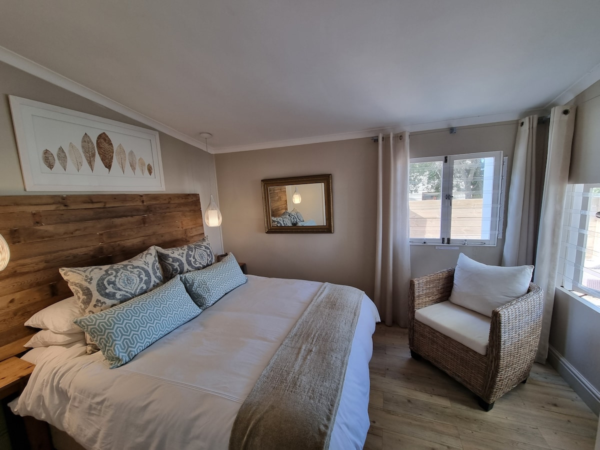 A cozy bedroom features a spacious bed adorned with decorative cushions and a light, neutral bedspread. Natural light enters through two windows, illuminating the room. A wicker chair complements the wooden elements, and a mirror adds depth to the space.
