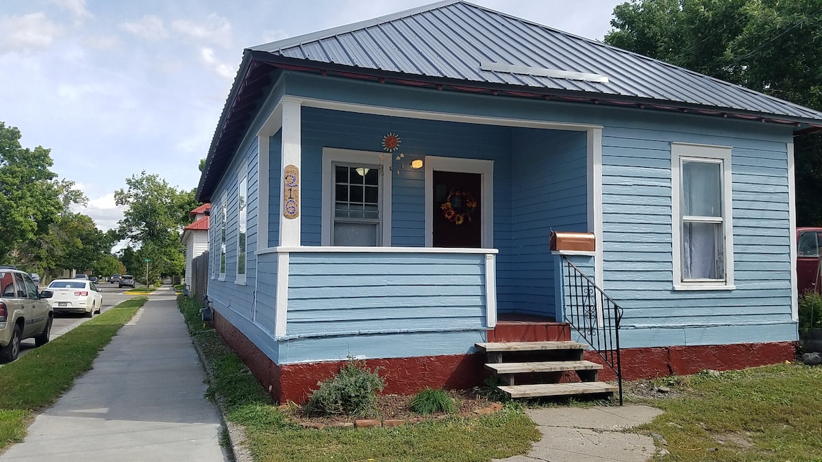 The exterior of a charming blue railroad house presents a welcoming porch with steps leading to the entrance. Large windows allow natural light to filter in, and the well-maintained yard includes greenery along the sidewalk. The property features a sloped roof and decorative details.