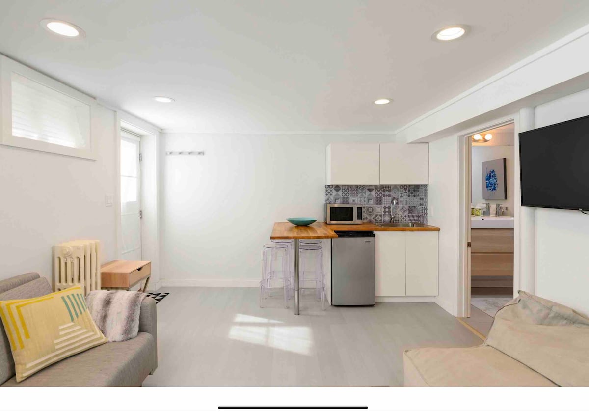 A bright, newly remodeled basement unit features a kitchenette with modern appliances, including a refrigerator and microwave. A comfortable seating area with a sofa bed is adjacent to a dining space highlighted by bar stools. Natural light enters through a window, enhancing the open layout.
