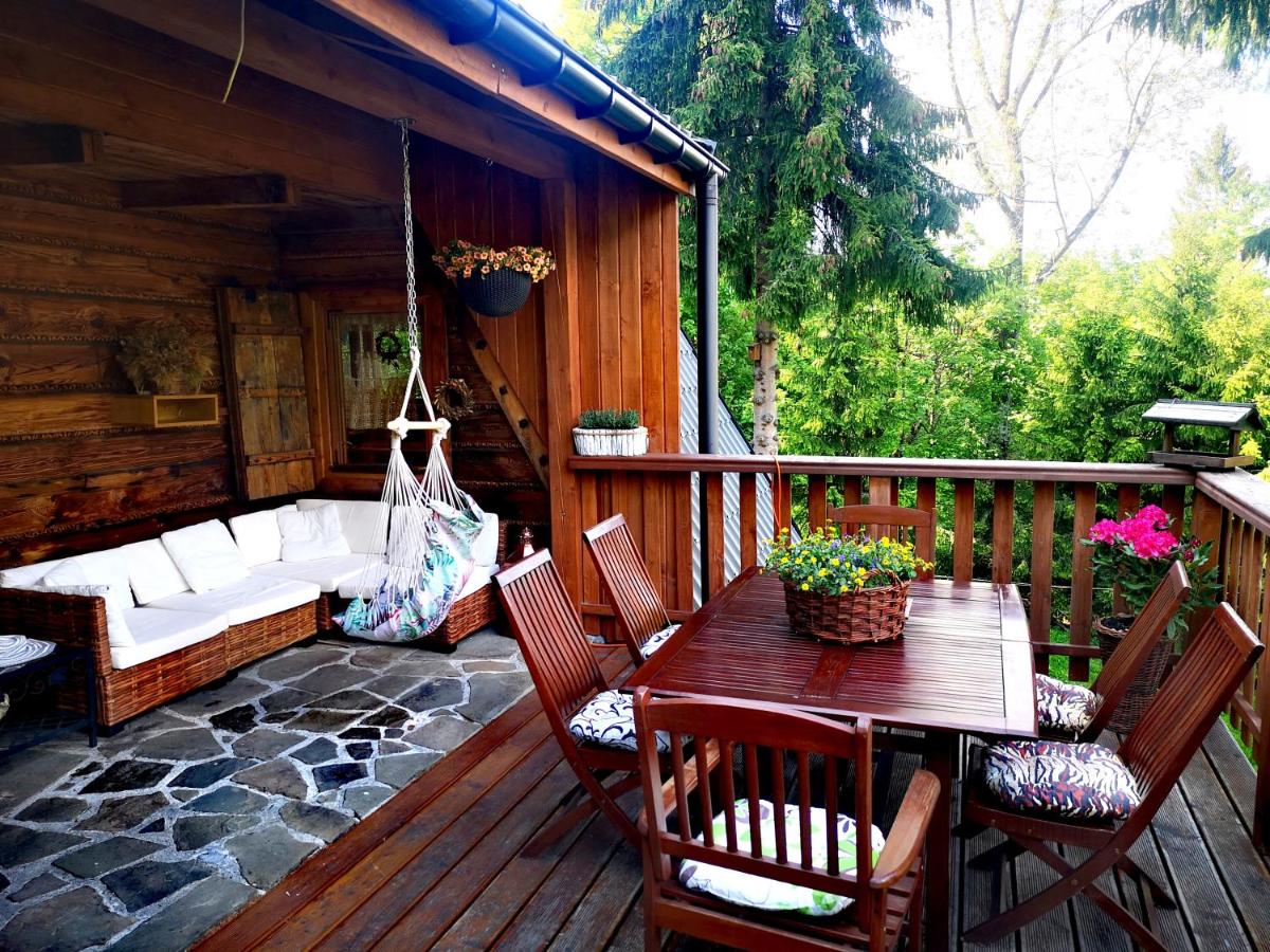 An outdoor terrace features a wooden dining table surrounded by chairs, with a cozy seating area including a suspended swing chair. Potted plants add vibrant greenery, while natural stone flooring complements the rustic wooden walls and surrounding lush foliage.