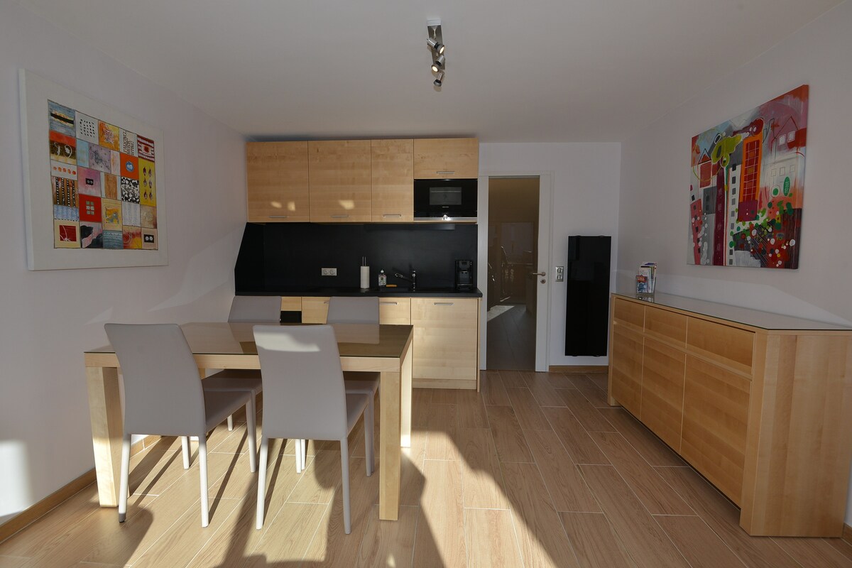 A modern kitchenette is showcased, featuring light wooden cabinetry, a dark countertop, and appliances including a microwave and coffee maker. A dining table with two chairs is present, alongside vibrant artwork adorning the walls. Natural light fills the space, enhancing its inviting simplicity.