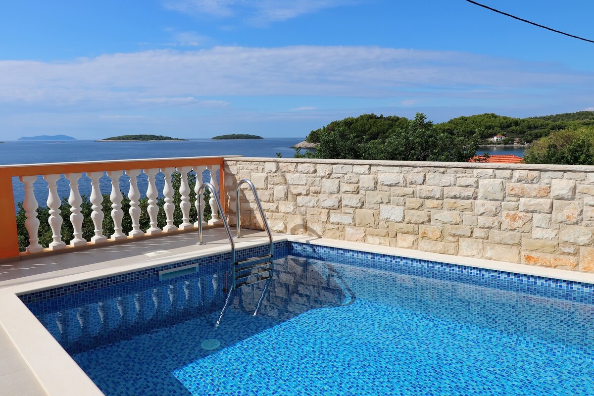 A private pool is set against a backdrop of the sea, featuring clear blue water and steps leading down into it. The surrounding stone wall offers privacy, while a view of the horizon and distant islands can be seen under a bright blue sky.