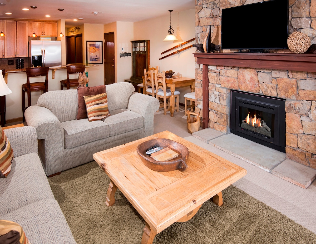 A cozy living area features a stone fireplace with a mounted television above. The room includes a light-colored sofa adorned with striped pillows, complemented by a wooden coffee table and a plush area rug. The dining area is visible in the background, showcasing a wooden table with chairs.
