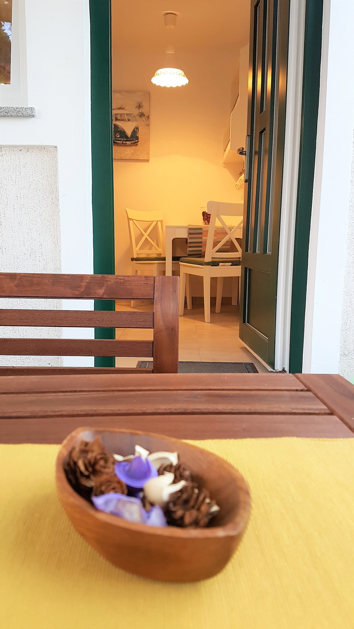 A wooden bowl filled with pine cones and purple flowers is placed on a yellow tablecloth. In the background, a door reveals a glimpse of a dining area furnished with white chairs and a calm, warm light illuminating the space.