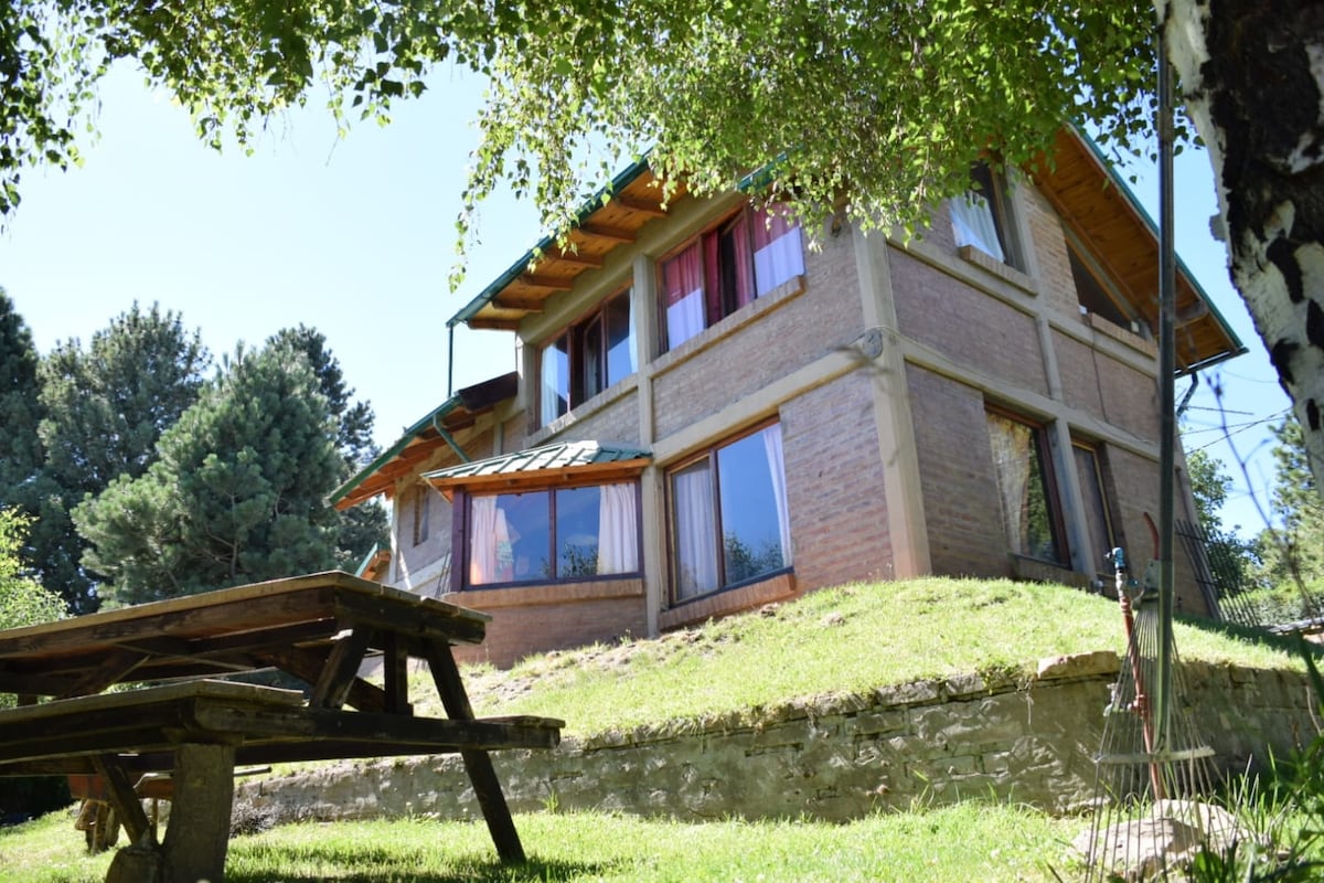 The exterior of a spacious multi-level cabin is shown, featuring large windows that allow for abundant natural light. A wooden picnic table is positioned on the green lawn, surrounded by trees and shrubs, creating a peaceful outdoor setting.