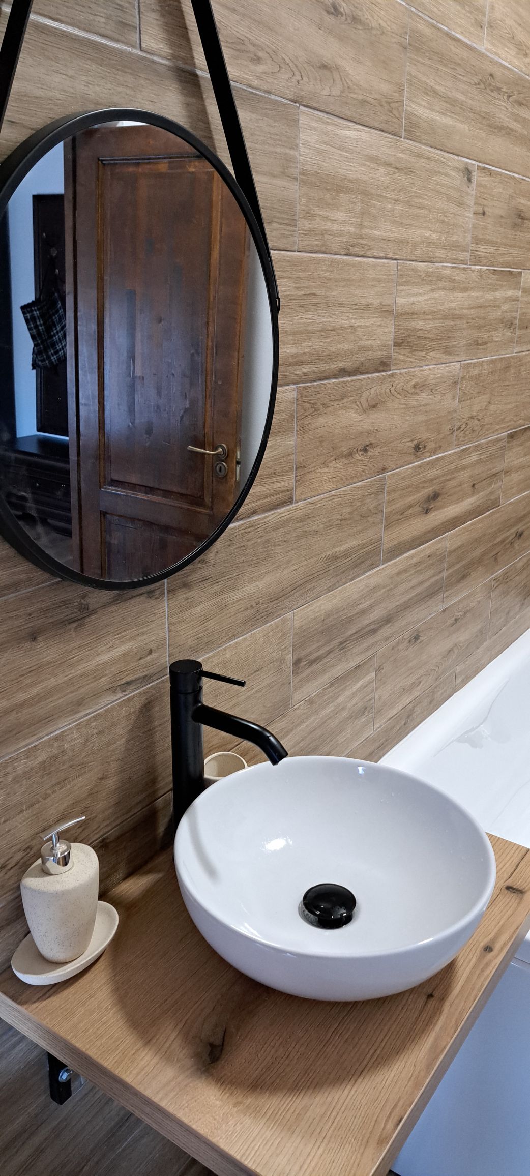 A modern bathroom features wooden wall panels and a round mirror above a vessel sink. A minimalistic faucet is mounted beside the sink, which rests on a wooden countertop. A creamy-colored soap dispenser is also present on the counter, complementing the design.