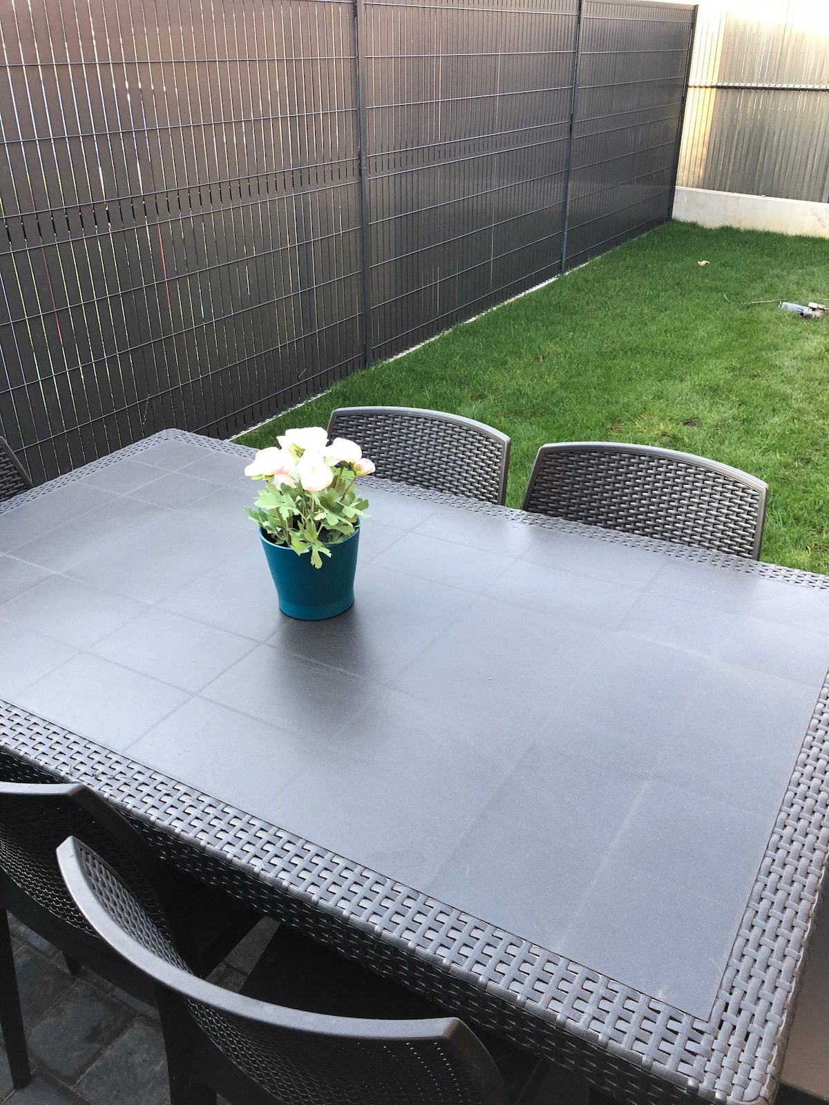 An outdoor dining area is showcased with a large rectangular table surrounded by four chairs. A blue pot containing a small flower arrangement is centered on the table. The background features a neatly maintained grassy area, bordered by a modern black fence.