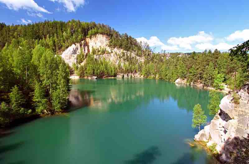 A tranquil lake surrounded by lush green trees and rocky cliffs reflects clear blue skies. The waters exhibit shades of turquoise, enhancing the natural beauty of the area.