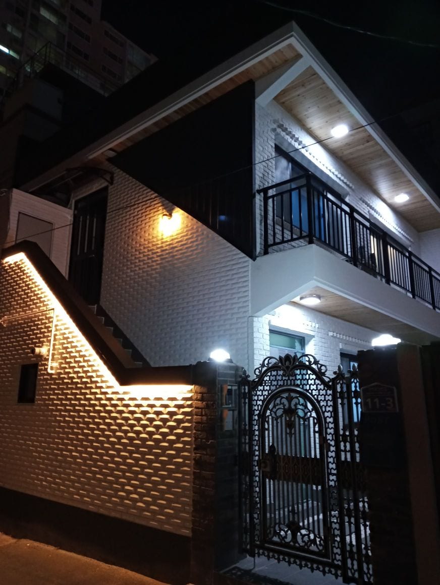 The exterior of the accommodation showcases a modern two-story building with textured white walls and decorative black accents. Soft lighting highlights the welcoming entrance and staircase, while a vintage-style gate adds charm to the property.
