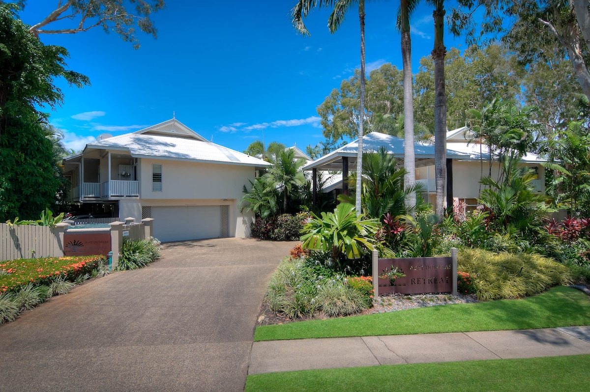 The exterior of Port Douglas Retreat is depicted, featuring well-maintained gardens with tropical plants. Two studio apartment buildings are visible, each with a traditional Australian design and a light-colored roof. A smooth driveway leads to the entrance, surrounded by lush greenery.