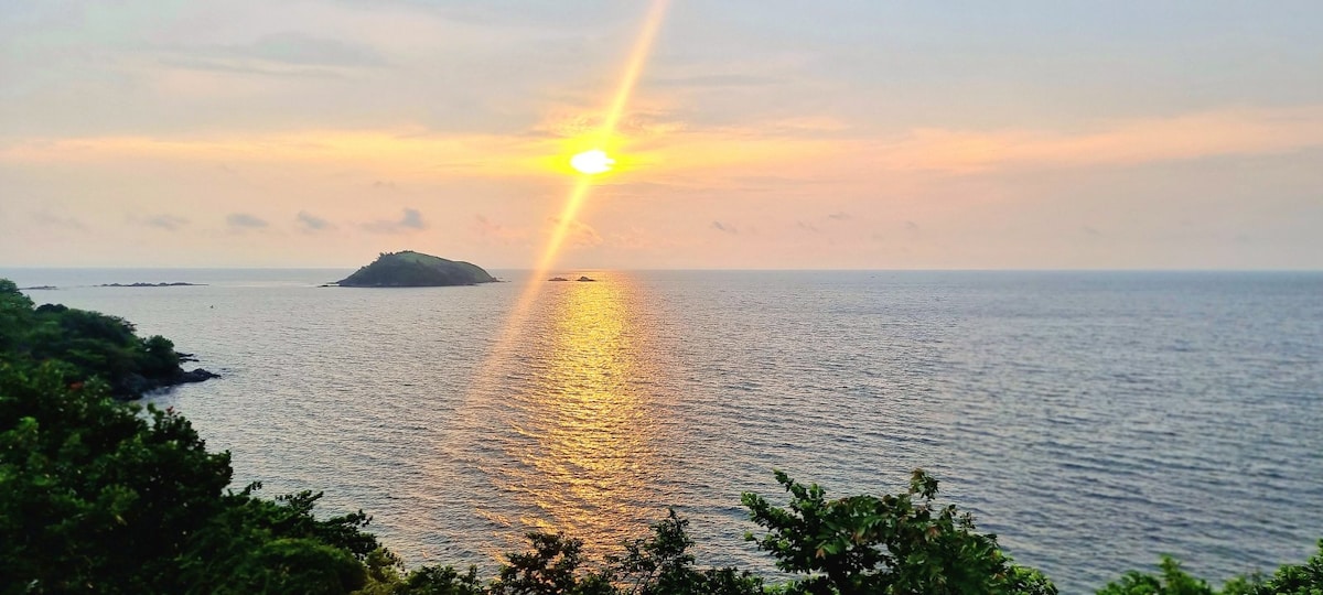 A serene view of the ocean at sunset, featuring calm waters reflecting golden hues. An island is visible in the distance, with lush greenery framing the scene, enhancing the connection to nature and tranquility.