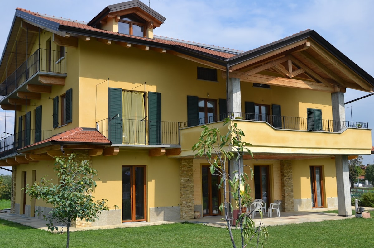 A modern, two-story building features a warm yellow façade complemented by wooden accents. Balconies and large windows allow natural light to flow throughout, while an inviting lawn area offers space for relaxation. The overall architecture showcases a blend of rustic charm and contemporary design.