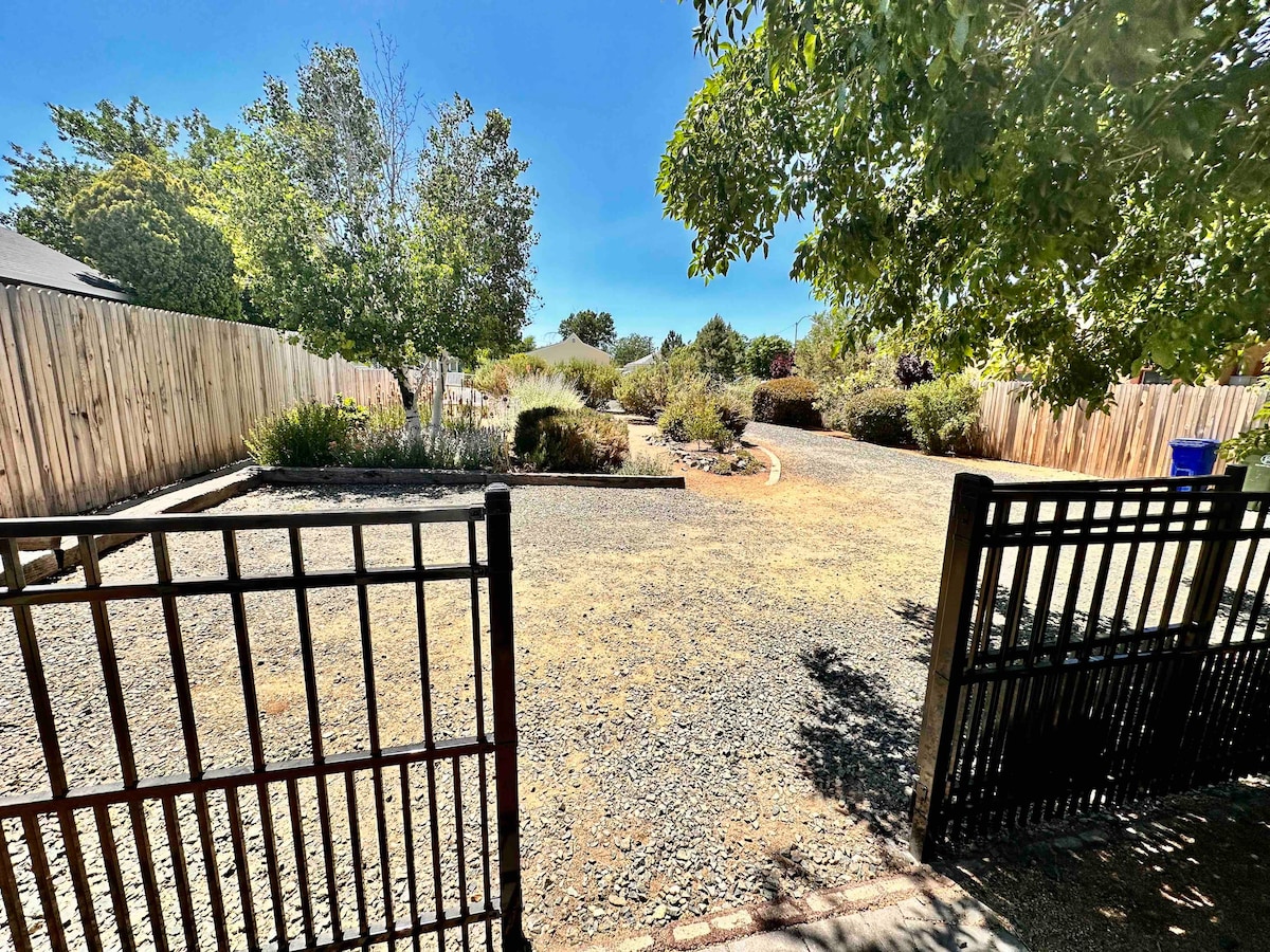 A gated entry leads into a spacious outdoor area, surrounded by wooden fencing and lush greenery. The gravel path curves through the landscape, bordered by a variety of plants and trees. Bright sunshine casts a warm light on the scene, enhancing the inviting atmosphere.