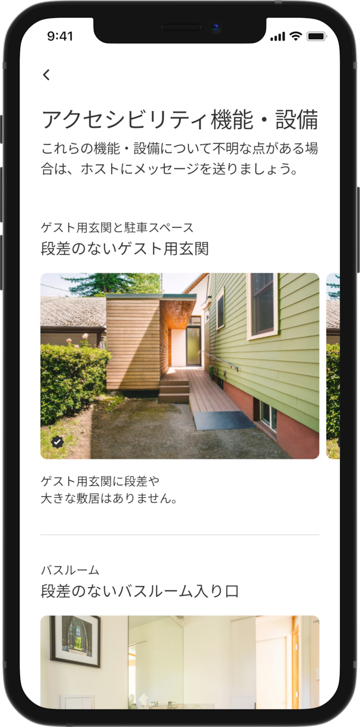 携帯電話にはAirbnbのリスティングの詳細情報が表示されています。メインの見出しには「アクセシビリティ機能・設備」とあり、「ゲスト用玄関と駐車場」や「バスルーム」のように機能・設備がグループ化されています。「ゲスト用玄関と駐車場」の下に「段差のないゲスト用玄関」という見出しと「ゲスト用玄関に段差や高さのある敷居はありません」という機能・設備の説明文があります。その下には、車椅子用スロープをはじめとするリスティングの設備・機能の画像スライドショーがあります。