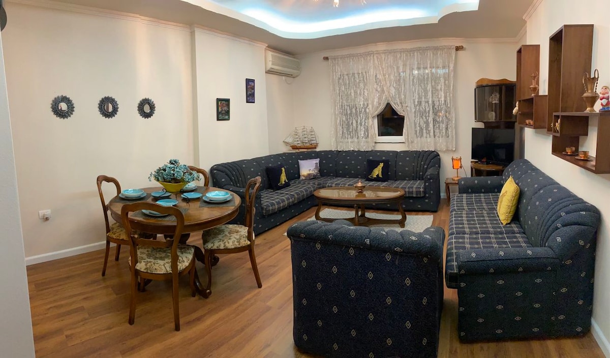 A spacious living area showcases a large blue patterned sectional couch arranged around a wooden coffee table. A round dining table, set for six, features decorative plates and a floral centerpiece. Natural light filters through sheer curtains above the window.