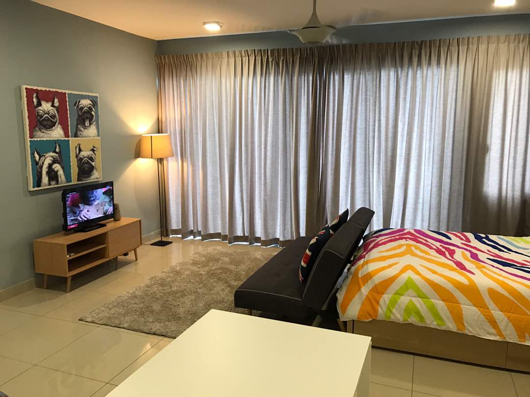 A cozy bedroom is furnished with a spacious bed featuring a colorful patterned blanket. A comfortable sofa is positioned next to a soft area rug. Natural light filters through floor-to-ceiling curtains, and a TV is mounted on a wooden media unit.