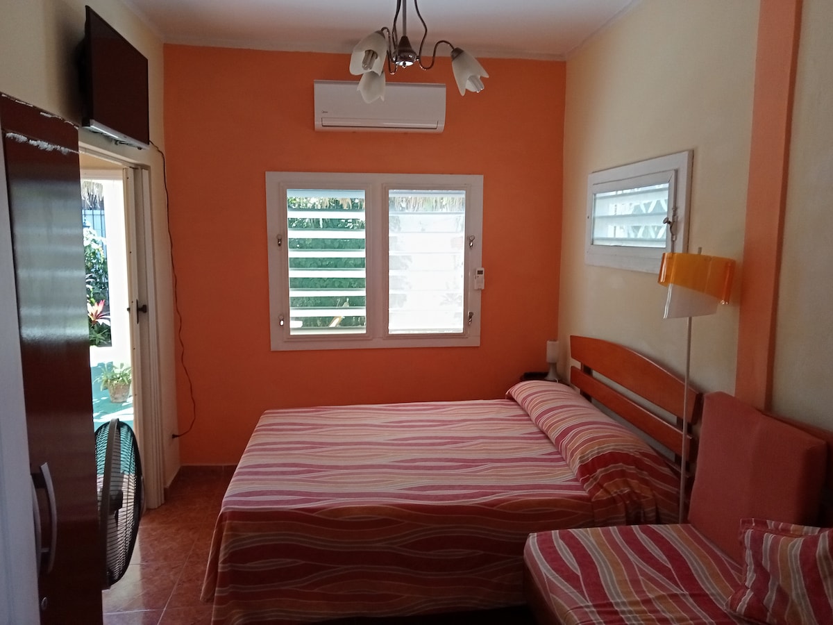 A light-colored room features an orange accent wall and is furnished with a striped bedspread covering a spacious bed. Two windows allow natural light to enter, and air conditioning is provided. A fan, television, and small sitting area are visible, enhancing comfort.