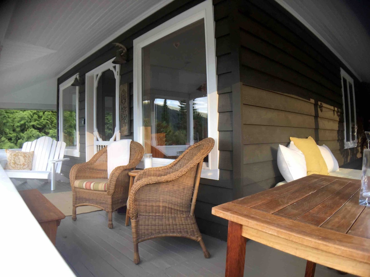 A private veranda is showcased, featuring two wicker chairs with soft cushions and a wooden table. The space is framed by expansive windows that provide views of the lush surroundings, promoting a peaceful atmosphere. Decorative elements enhance the inviting ambiance.