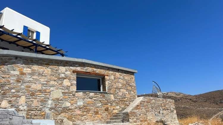 A stone structure with a curved wall features a large window providing views of the surrounding landscape. A staircase leads to the entrance, while a pergola offers shaded outdoor space. The clear blue sky enhances the building's modern design.