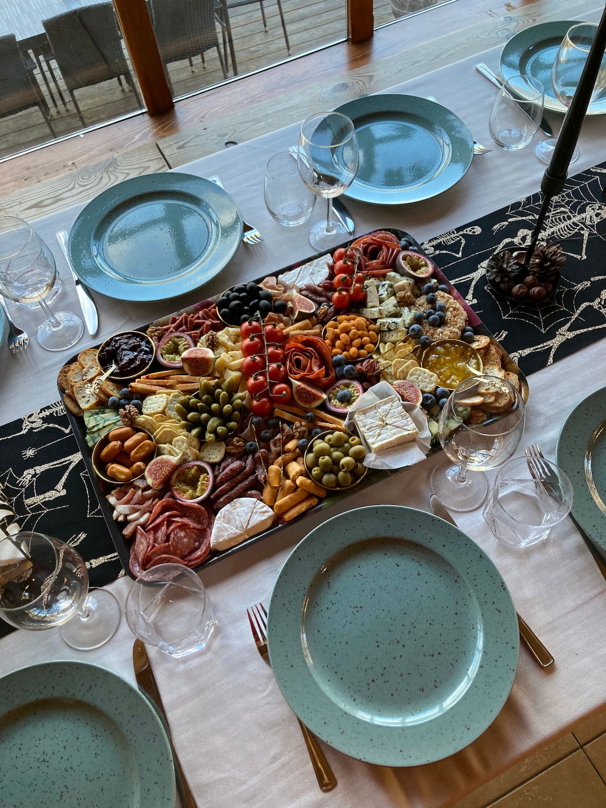 A large wooden platter is filled with an array of cheeses, meats, olives, fruits, and nuts. Surrounding the platter are several turquoise plates and elegant glassware, all set on a table adorned with a decorative runner featuring a nature-inspired design.