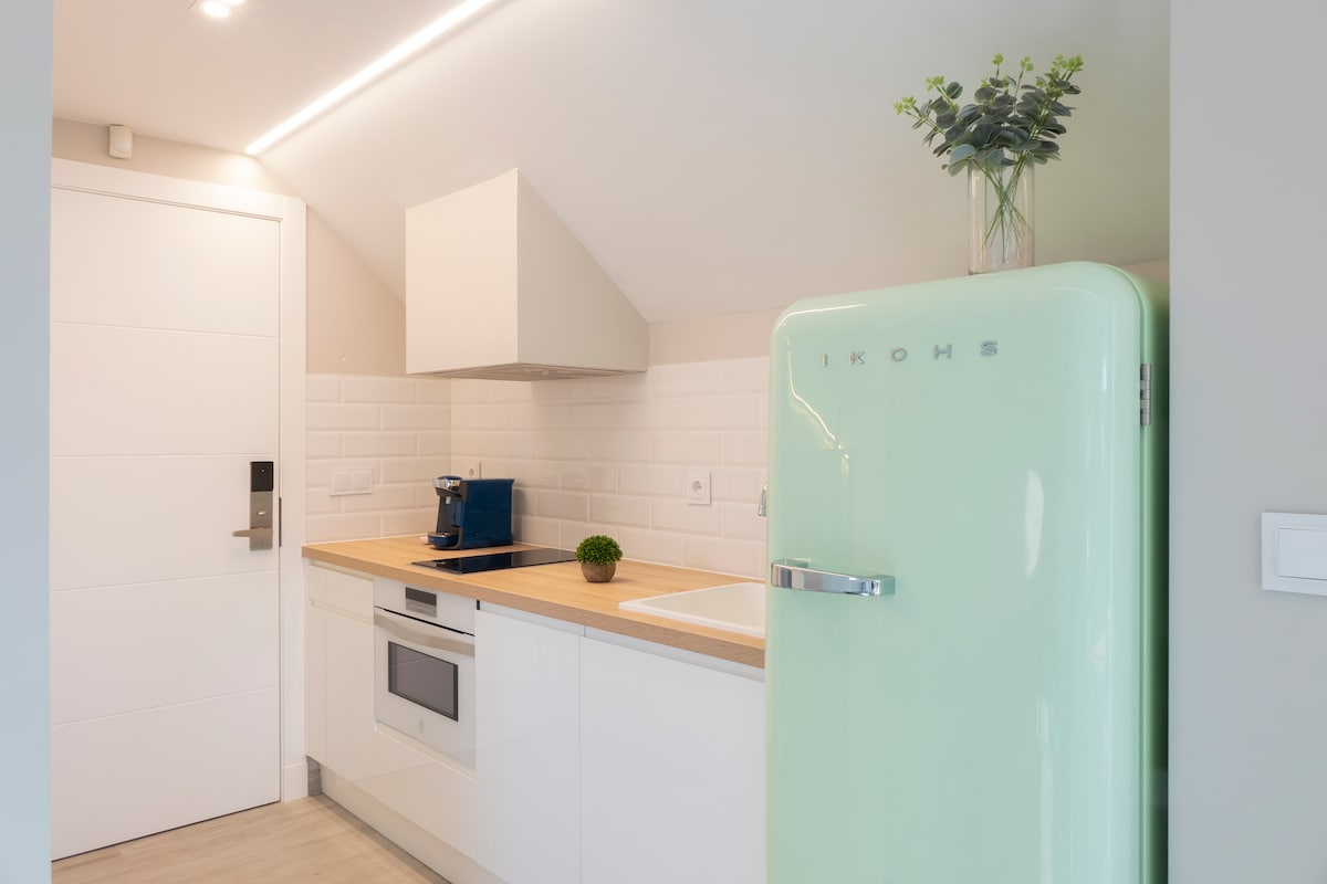 A modern kitchen is presented, featuring clean white cabinetry and light wooden countertops. A vintage-style mint-green refrigerator is prominently displayed. Essential appliances, including a stovetop and oven, are integrated into the design, complemented by minimal decorative elements like a small potted plant.
