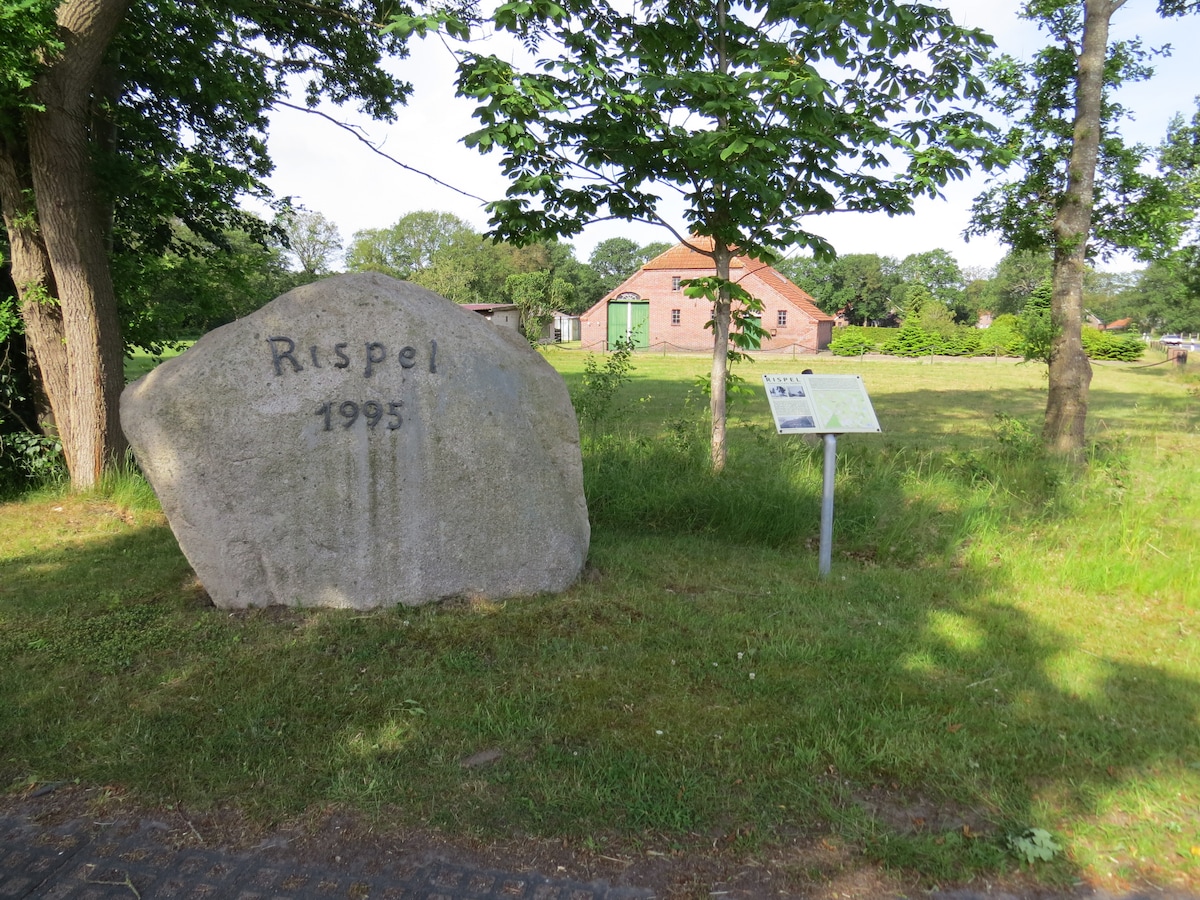 A large gray stone is visible, engraved with the word 'Rispel' and the year '1995.' Nearby, an informational sign stands on a grassy area. In the background, a rustic brick building is surrounded by trees and an expansive green landscape.