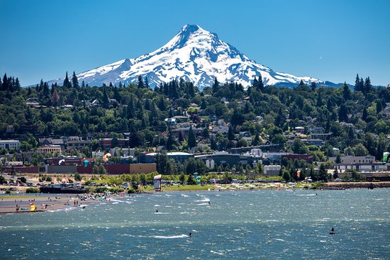 A vibrant scene showcases the majestic snow-capped Mount Hood rising above lush green hills, while clear blue skies offer a bright backdrop. The foreground features a lively beach area with various water sports activities, illustrating the natural beauty of the Columbia River Gorge.