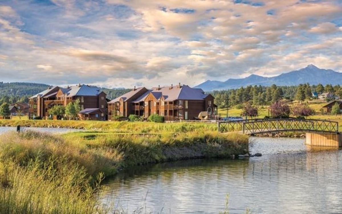 A serene landscape showcases a cluster of wooden buildings alongside a reflective body of water, framed by lush greenery and distant mountains. A wooden footbridge crosses the water, inviting visitors to explore the natural surroundings under a sky adorned with soft clouds.