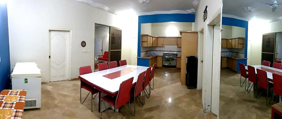 A spacious dining area features a large white table surrounded by red chairs. An open kitchen area is visible in the background, with wooden cabinetry and modern appliances, including a stove and refrigerator. A side door leads to another room, enhancing accessibility.