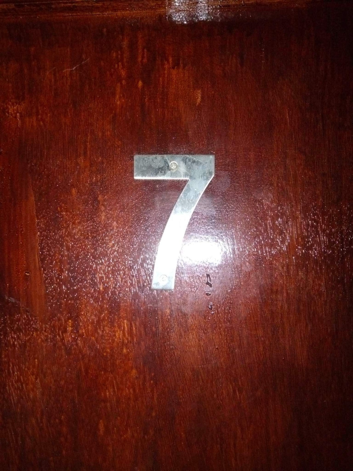 A close-up image captures a large metallic numeral '7' affixed to a polished wooden door. The rich wood grain is visible, displaying a deep brown hue with a glossy finish.