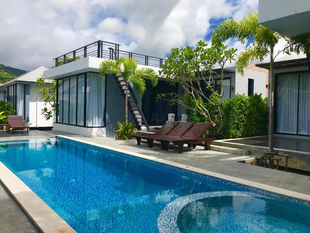 A shared swimming pool glimmers under a partly cloudy sky, flanked by modern villas featuring large glass windows. Lush tropical greenery adds a vibrant touch, while a wooden deck with lounge chairs invites relaxation. A staircase leads to an upper area, enhancing the dynamic architectural design.