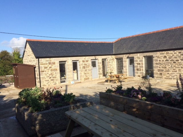 A well-maintained stone cottage exterior is shown under a clear blue sky. The entrance is marked by multiple grey doors, with a communal outdoor seating area visible. Nearby, a landscaped area features flowering plants, enhancing the inviting atmosphere of the shared space.