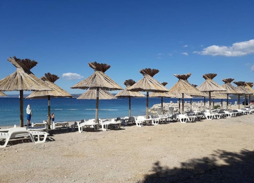 A sandy beach scene features several thatched umbrellas providing shade over a row of white lounge chairs. The clear blue water stretches into the distance, with gentle waves lapping at the shore. A few individuals can be seen enjoying the beach.