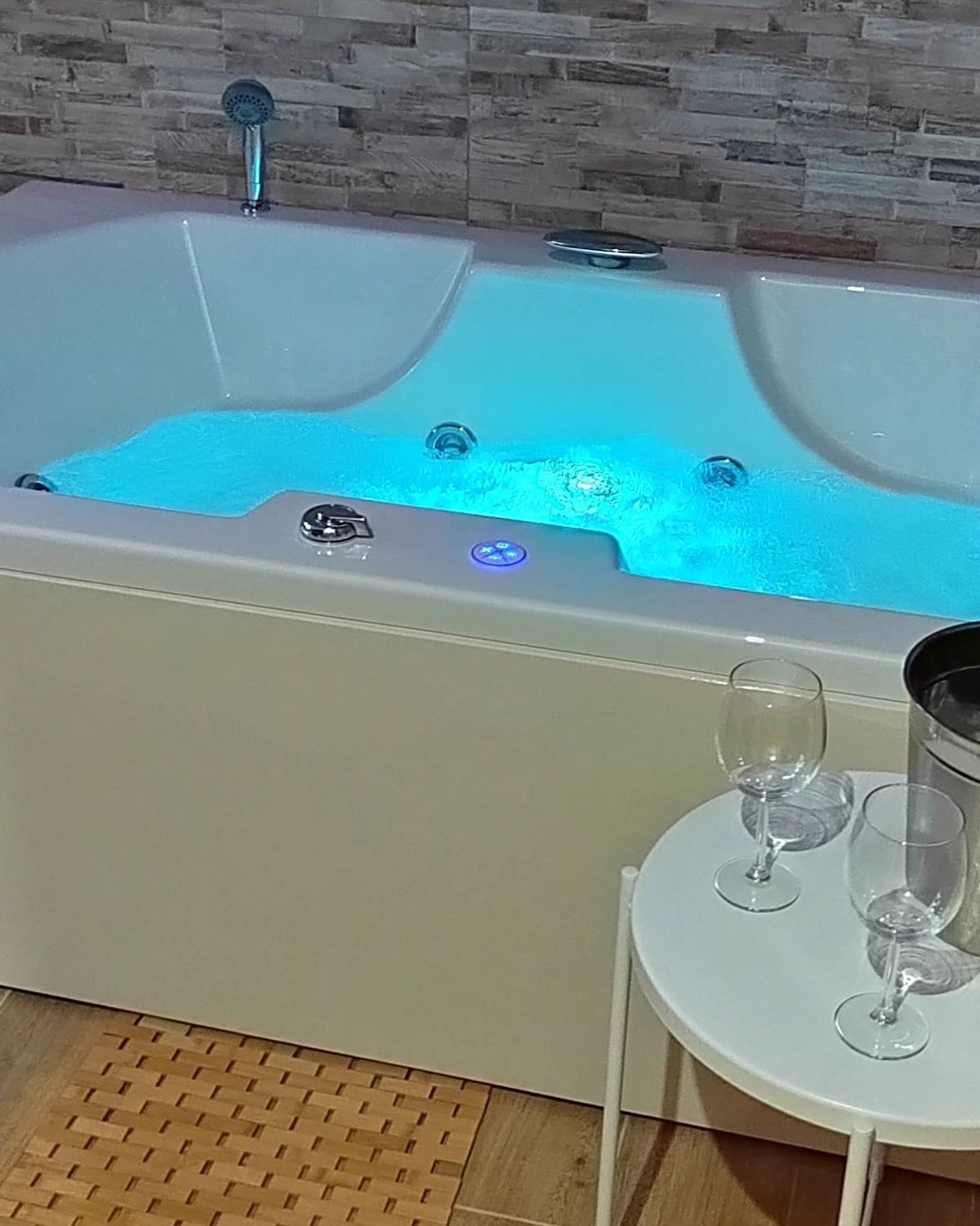 A modern jetted tub is featured, illuminated with soft blue lighting, enhancing the relaxation experience. Nearby, a small table holds two champagne glasses and a bucket, set against a backdrop of wooden flooring and textured wall tiles.