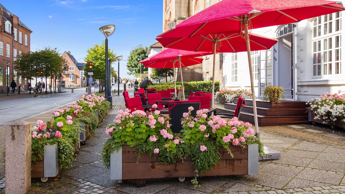 An outdoor seating area is shown with red umbrellas and seating arrangements. Colorful flower boxes filled with blooming plants line the edge, and a tree-lined street is visible in the background, contributing to the inviting ambiance of the space.