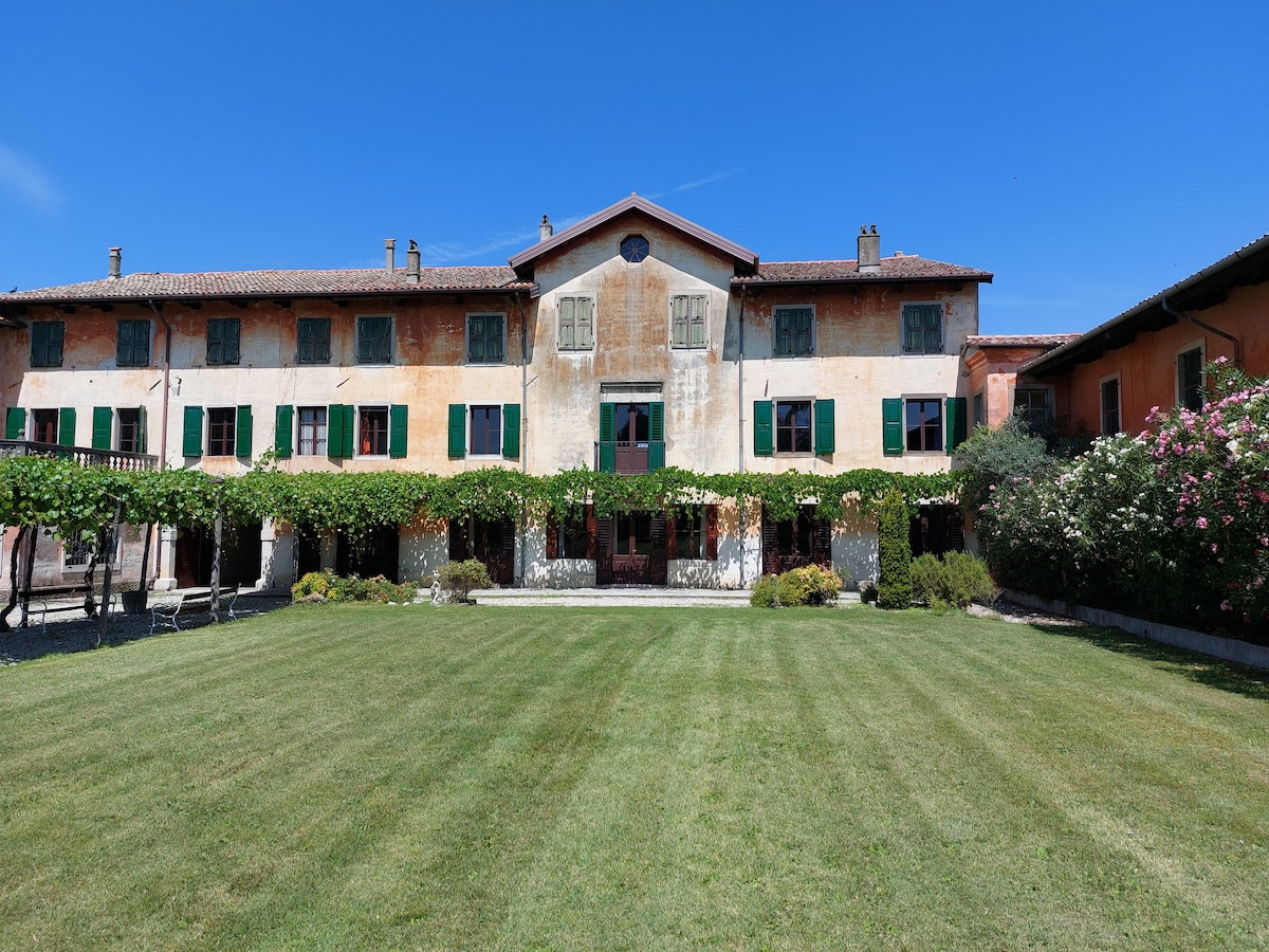 The villa's charming exterior is highlighted by a weathered facade, framed by green shutters. A well-maintained lawn leads to a covered terrace adorned with climbing vines. The structure features multiple windows and balconies, providing a glimpse of the surrounding park and gardens.