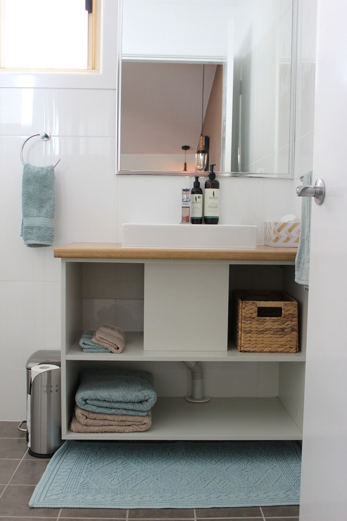 A modern bathroom is displayed, featuring a wooden countertop with a square sink. A large mirror reflects the space, while neatly arranged toiletries rest alongside. Beneath the countertop, a shelf holds rolled towels and a woven basket, complemented by a small rug on the grey tiles.