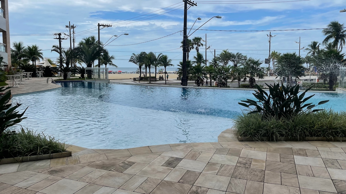 A scenic pool area is depicted, featuring a clear blue water surface that reflects the sky. Surrounding palm trees and landscaped greenery create a tropical ambiance. The beach is visible in the distance, just beyond the pool area.