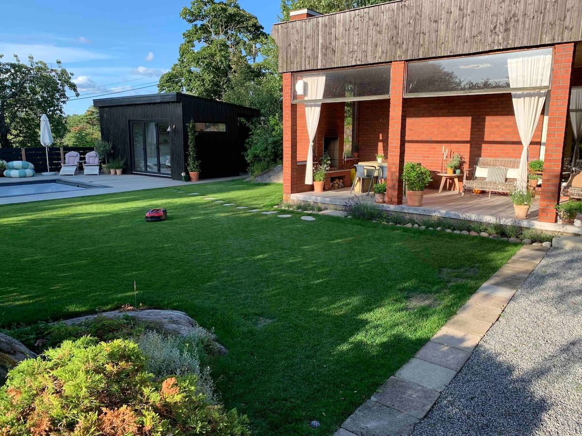 A spacious garden is seen with well-maintained grass that transitions to a light gravel path. The house features large windows, providing a view of the interior and connecting outdoor and indoor spaces. A pool area is visible to the left, with seating arrangements nearby.