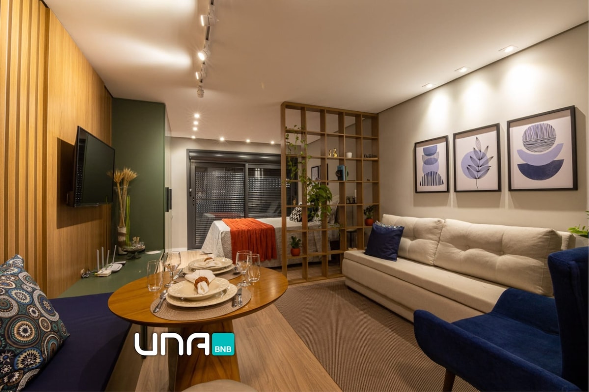 The living area features a light-colored sofa paired with a blue chair, accompanied by a wooden dining table set for four. A shelf with plants adds greenery, while three framed artworks decorate the wall, enhancing the space's calm aesthetic.