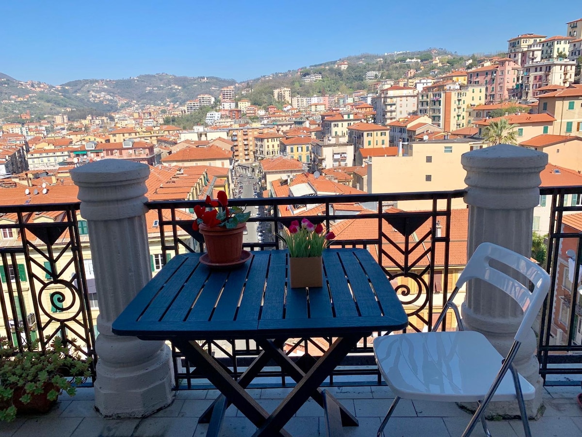 A balcony table with two chairs is positioned overlooking the cityscape. Potted flowers add a touch of color, while rooftops and hills are visible in the background, creating a scenic view of the historic center.