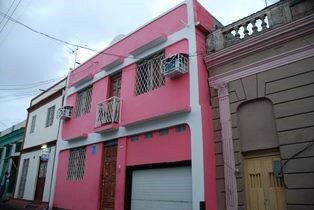 A two-story building in bright pink features decorative metal grilles on windows and a deep brown door. The structure is flanked by neighboring buildings in muted tones, emphasizing its vibrant color. An open garage is visible on the ground level.