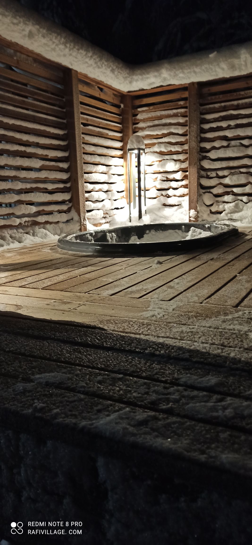 A warm wooden deck is featured, surrounding a round hot tub that glistens in the evening light. Snow-covered walls create a cozy enclosure, while a lantern casts a soft glow, enhancing the tranquil atmosphere.